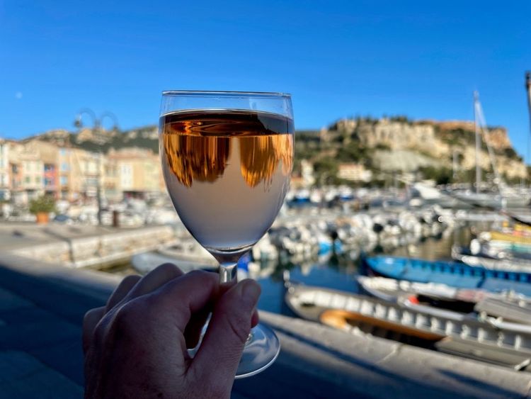 Mano femminile che regge un bicchiere di vino rosato, porto e centro storico di Cassis sullo sfondo